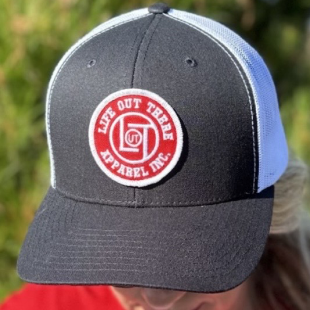 A person wearing the LOT Curved Bill Snapback Trucker Hat in black and white, featuring a red circular patch that reads Life Out There Apparel. The background is blurred greenery.