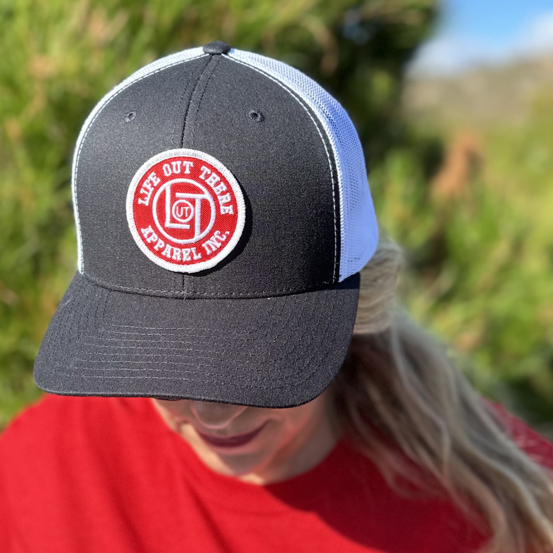 An individual is wearing the LOT Curved Bill Snapback Trucker Hat in black and white, featuring a red circular patch that reads Life Out There Apparel. The hat's curved bill enhances their style, perfectly paired with a red shirt against a backdrop of blurred greenery.