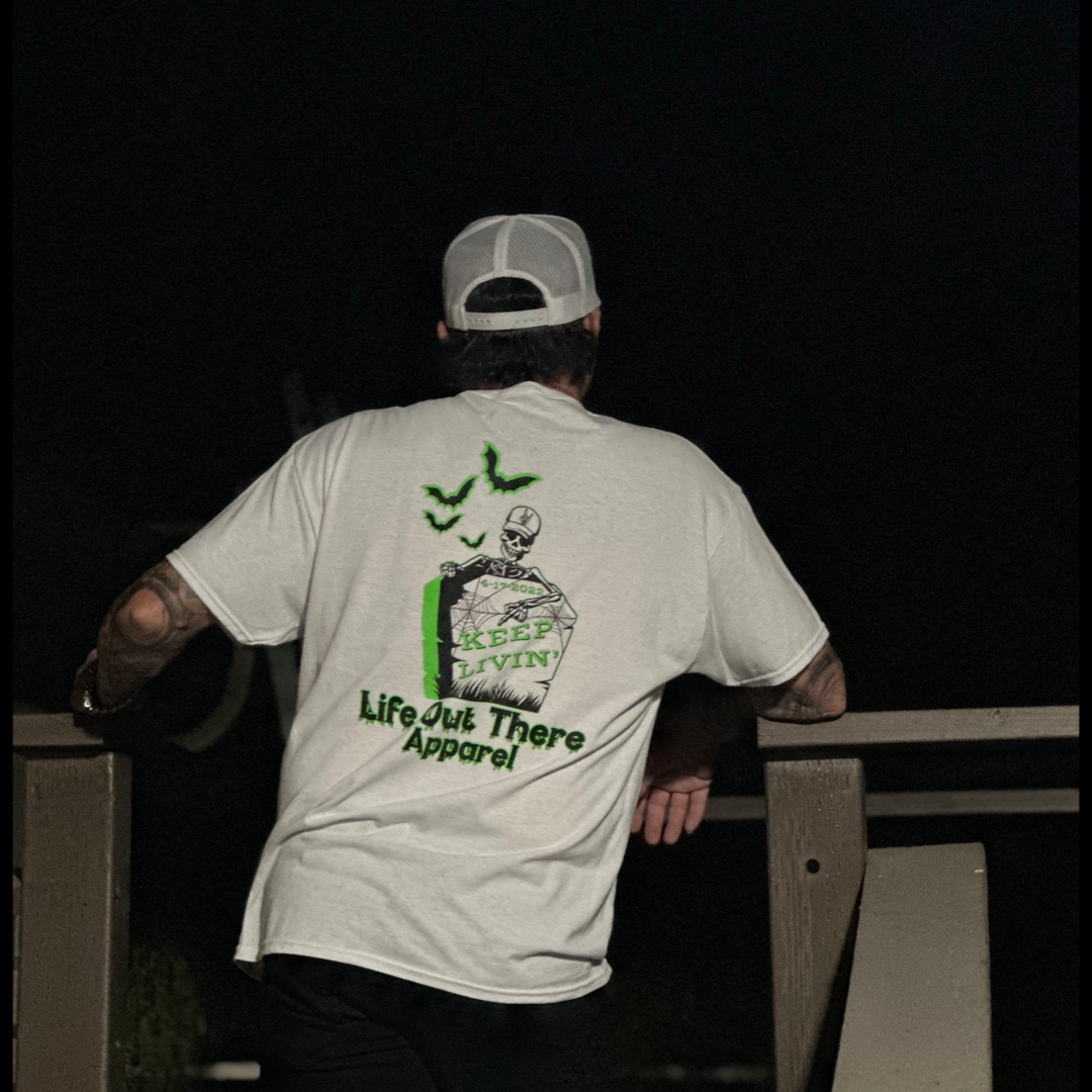 A person wearing the Unisex Keep Livin' T Shirt by Life Out There Apparel leans on a wooden railing. The back of the white shirt features an eerie Halloween-inspired design with skeletons, bats, and the phrases "Keep Livin" and "Life Out There Apparel," enhancing the spooky holiday vibe.