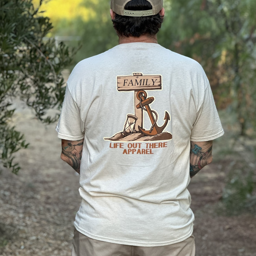 A man dons the Unisex Time Flies T Shirt in the sandstone color, showcasing a design with a wooden sign, rope, and anchor embellished with the words "FAMILY" and "LIFE OUT THERE APPAREL." He stands outside against a backdrop of blurred greenery, embodying the timeless message that time flies when cherished with loved ones.