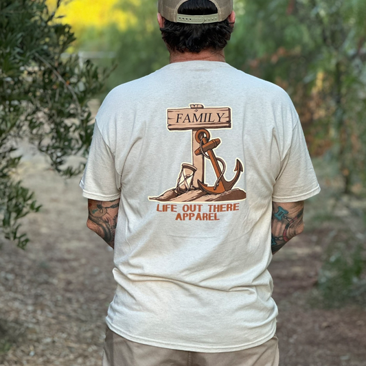 A person stands outdoors wearing the Life Out There Apparel Time Flies T Shirt in sandstone, featuring an anchor family design with “FAMILY” on a sign and “LIFE OUT THERE APPAREL” printed below the image on the back.