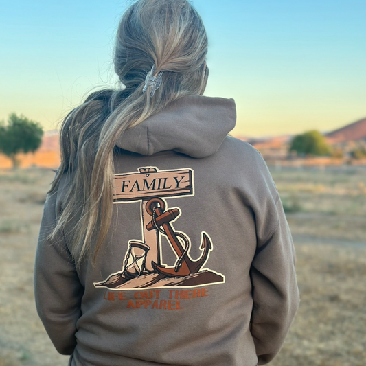A person with long blonde hair in a ponytail stands outdoors in a dry landscape, wearing the Life Out There Apparel Time Flies Hoodie - Safari, featuring a family anchor design and a "Family" sign for a warm, heartfelt vibe.