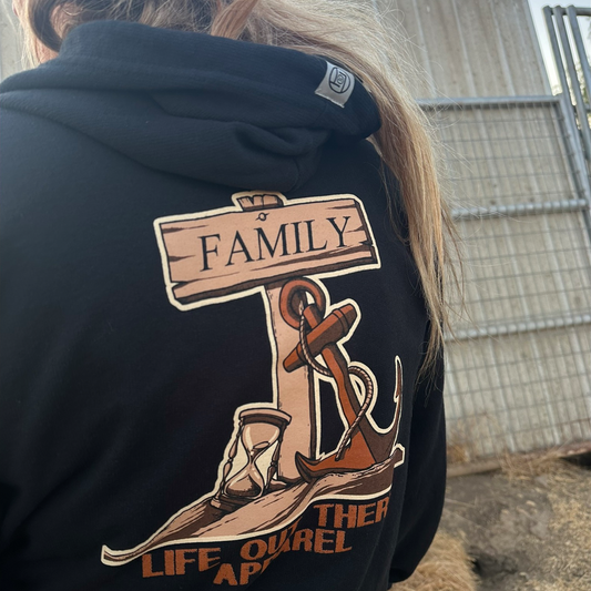A person with long hair is wearing the Unisex Time Flies "Zip-Up" Hoodie in black by Life Out There Apparel, featuring a design with an anchor and a wooden sign reading "Family," along with the phrase "Together Life OurEL Apis and Time Flies." In the background, there's a metal fence and part of a building.