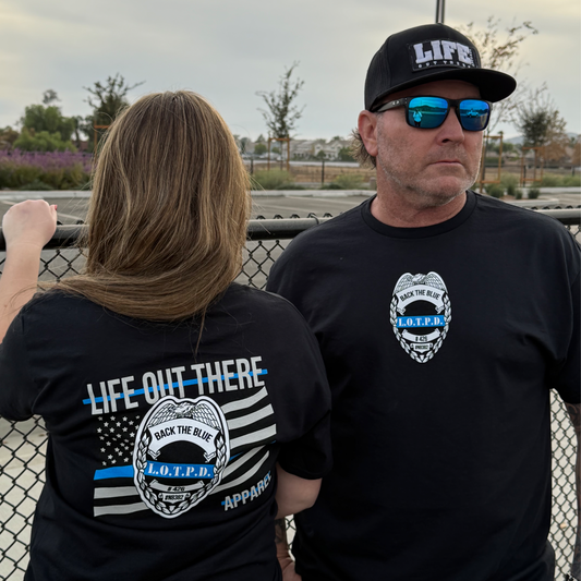 A man and woman stand by a fence in unisex black Back The Blue tees from Life Out There Apparel, each showcasing blue line law enforcement themes. The womans shirt includes a Sheriff’s Department emblem, while the man wears a Life branded cap.