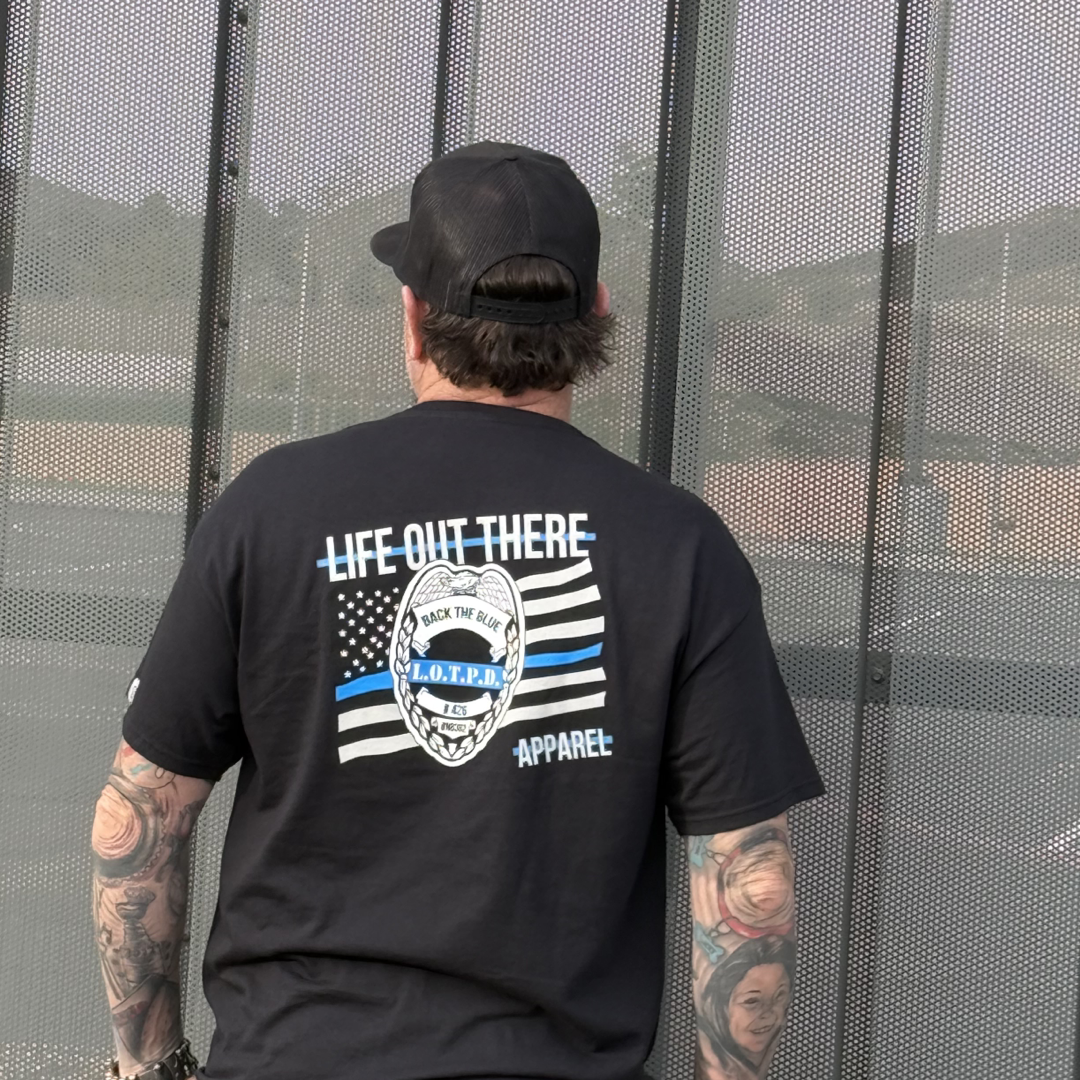 A tattooed individual in a black cap and Life Out There Apparels Back The Blue T-shirt, featuring Life Out There and a flag design on the back, stands solemnly facing a metal fence, subtly showing their support.