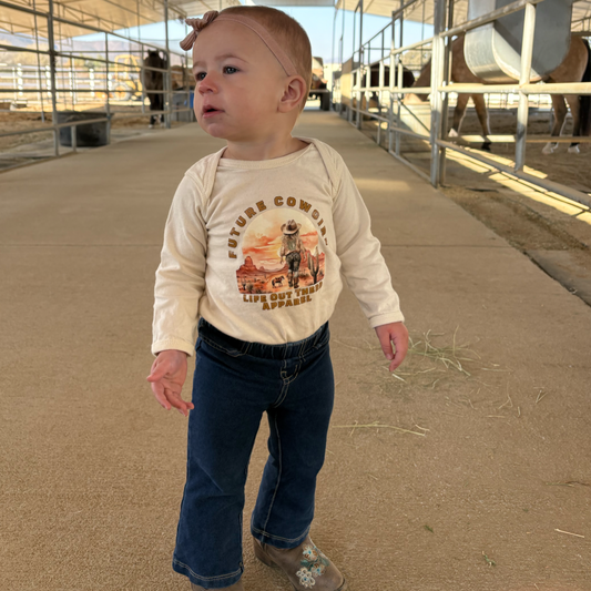 In a stable, a toddler wearing Life Out There Apparels Girls - Infant Future Cowgirl Snap Tee in beige stands curiously on the path surrounded by horses. She pairs the shirt with jeans and brown boots.