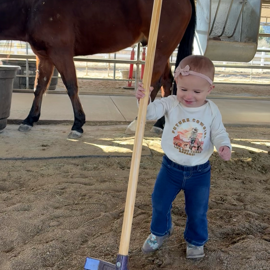 A child in a Future Cowgirl tee by Life Out There Apparel smiles with a wooden tool, wearing jeans and a pink headband. A horse is nearby as she stands on sandy ground under a canopy, with soft country music playing.