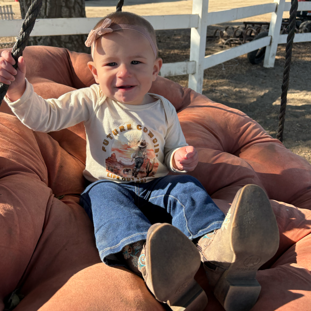 A baby sits on a cushioned swing outdoors wearing Life Out There Apparels Girls - Infant Future Cowgirl tee in natural beige. She pairs it with blue jeans, boots, and a head bow. Smiling, she grips the ropes with a wooden fence and trees as the picturesque country backdrop.