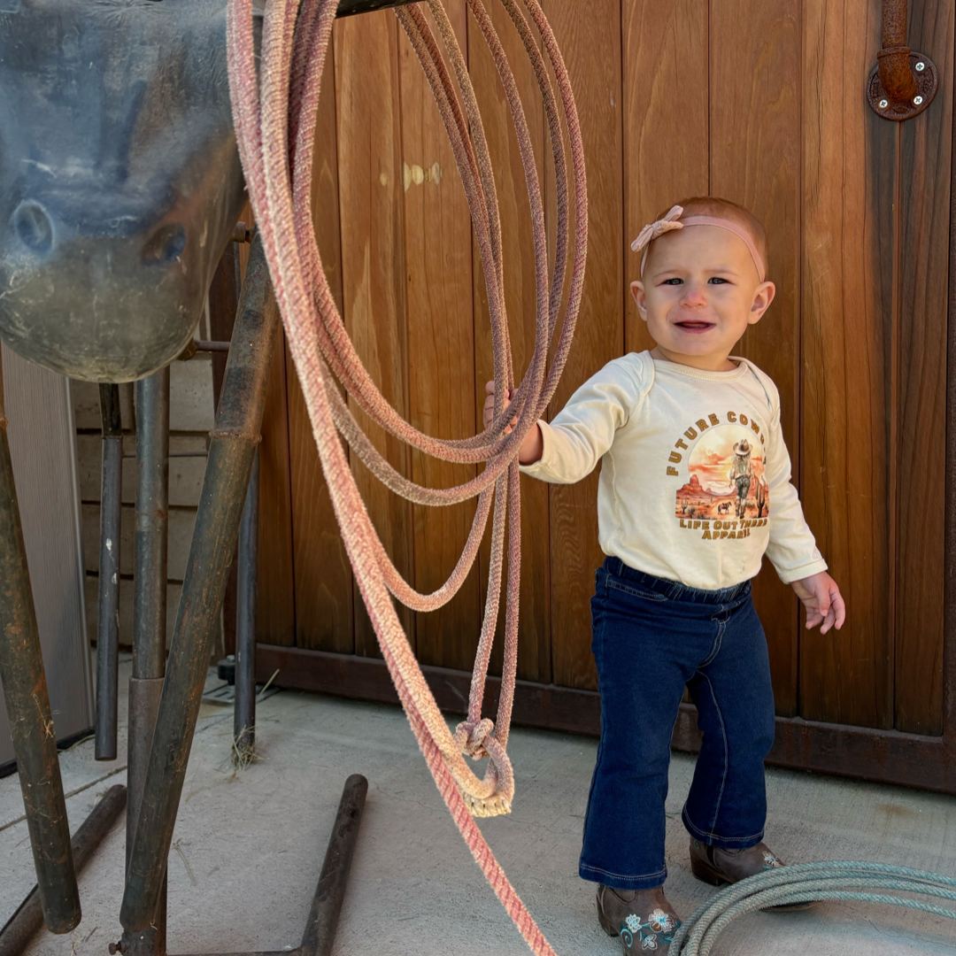 A toddler channels a future cowgirl by standing next to a metal horse structure, wearing her Infant Future Cowgirl snap tee from Life Out There Apparel, paired with jeans and boots. With a coiled rope in hand and a bow in her hair, she stands against the rustic charm of a wooden door.