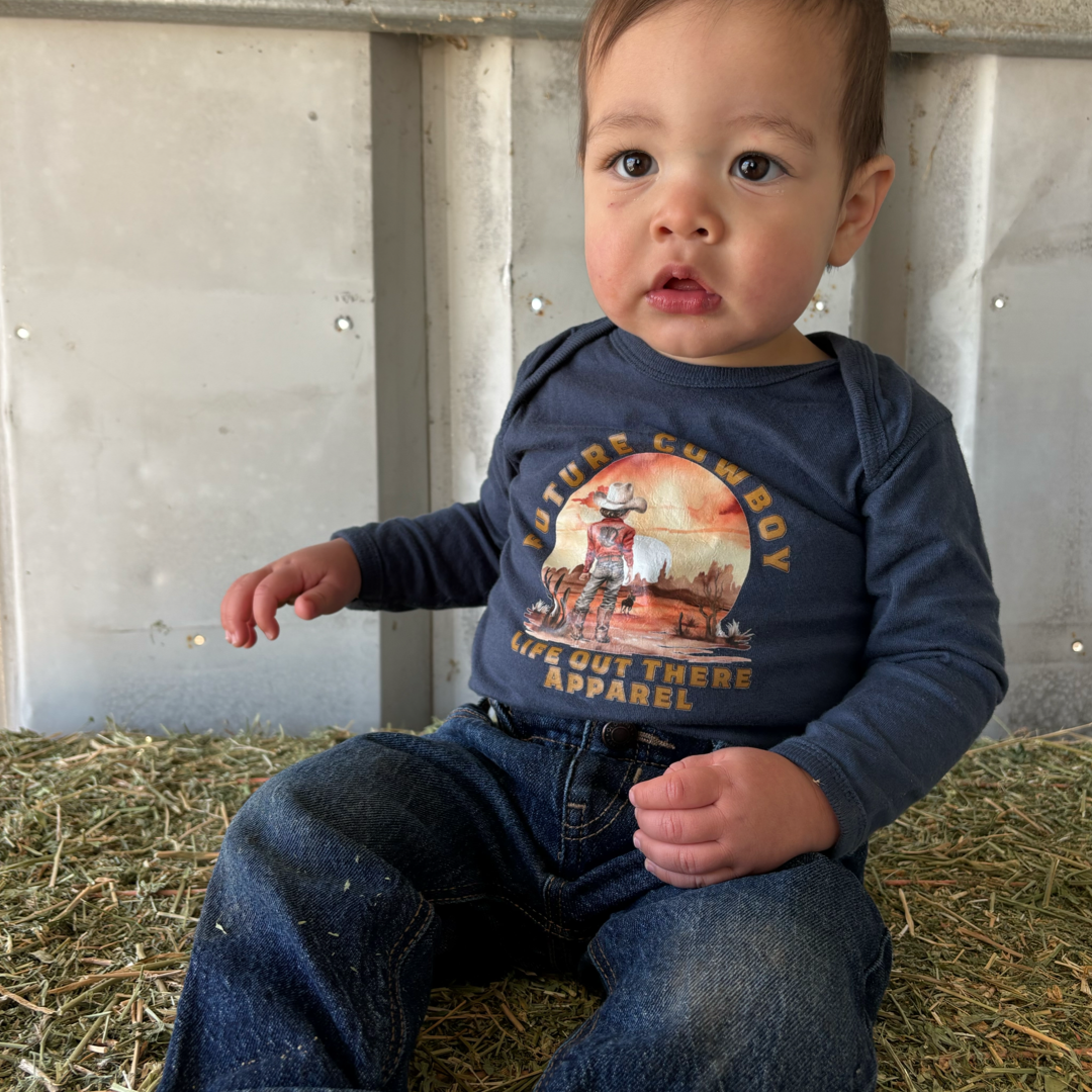 A toddler sitting on hay wears Life Out There Apparels Future Cowboy navy snap tee made of 100% cotton and denim jeans. The metallic wall frames their curious expression, with their left hand gently resting on the hay.