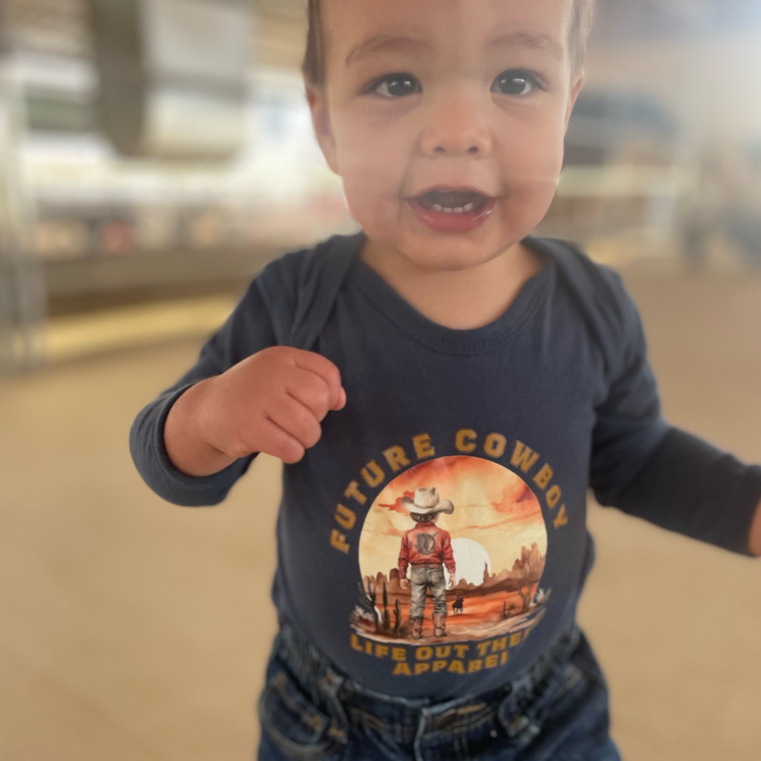 A toddler beams in a Life Out There Apparel shirt, clad in Denim Blue with Future Cowboy text and a cowboy design. The blurred background accentuates the childs joyful expression.