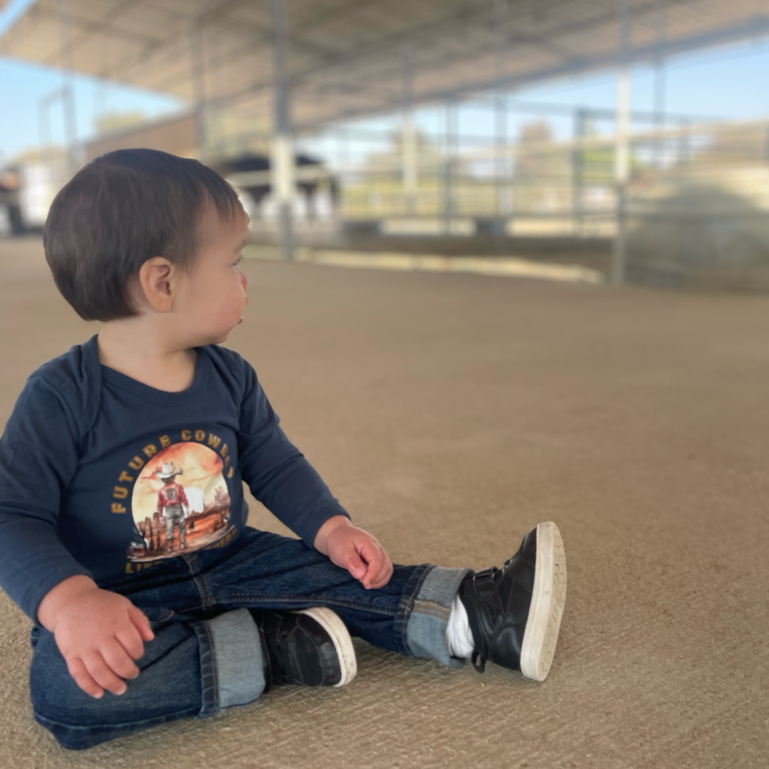 In a barn, a toddler sits on the concrete floor wearing Life Out There Apparels Boys Infant - Future Cowboy snap tee in denim. With short dark hair and side-gazing eyes, he is surrounded by the spacious barns fence and blurred animals in the background.