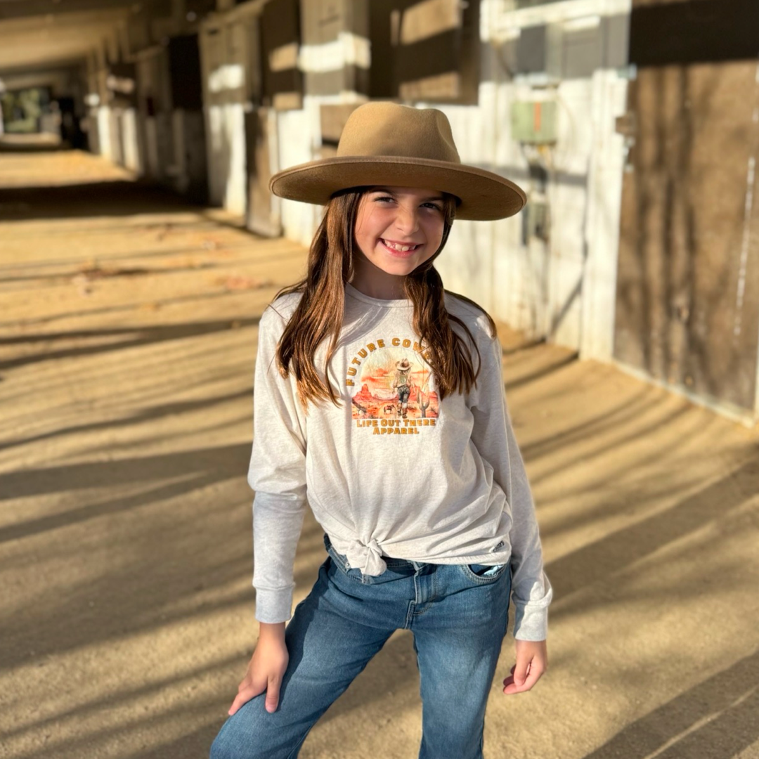 In a sunlit barn aisle, a young girl smiles wearing a Life Out There Apparel Future Cowgirl long-sleeve shirt and blue jeans. Her wide-brimmed hat casts shadows on the ground, echoing the timeless rhythms of country music.