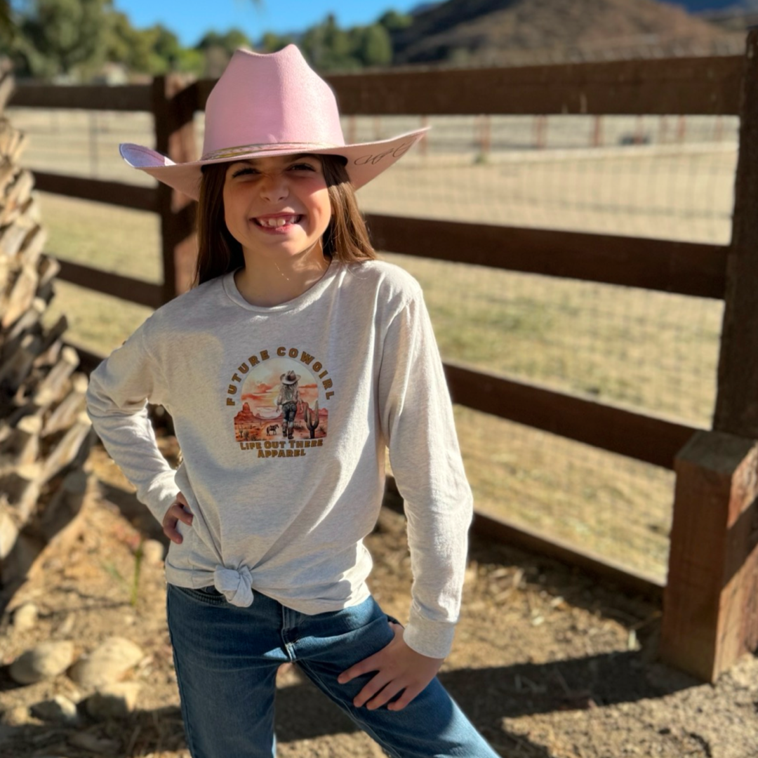 A young girl at a ranch beams with a hand on her hip, wearing a light pink cowboy hat and a Future Cowgirl long-sleeve shirt from Life Out There Apparel. Country music plays softly near a wooden fence and open field.