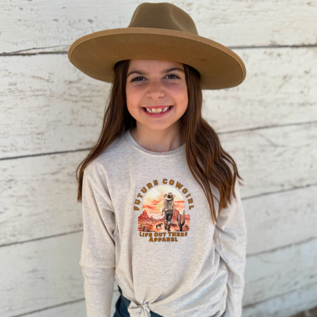 A smiling young girl in a Life Out There Apparel Future Cowgirl long-sleeve shirt, exuding country spirit, stands confidently before a white wooden wall. The shirt is available in natural beige, black, and red.