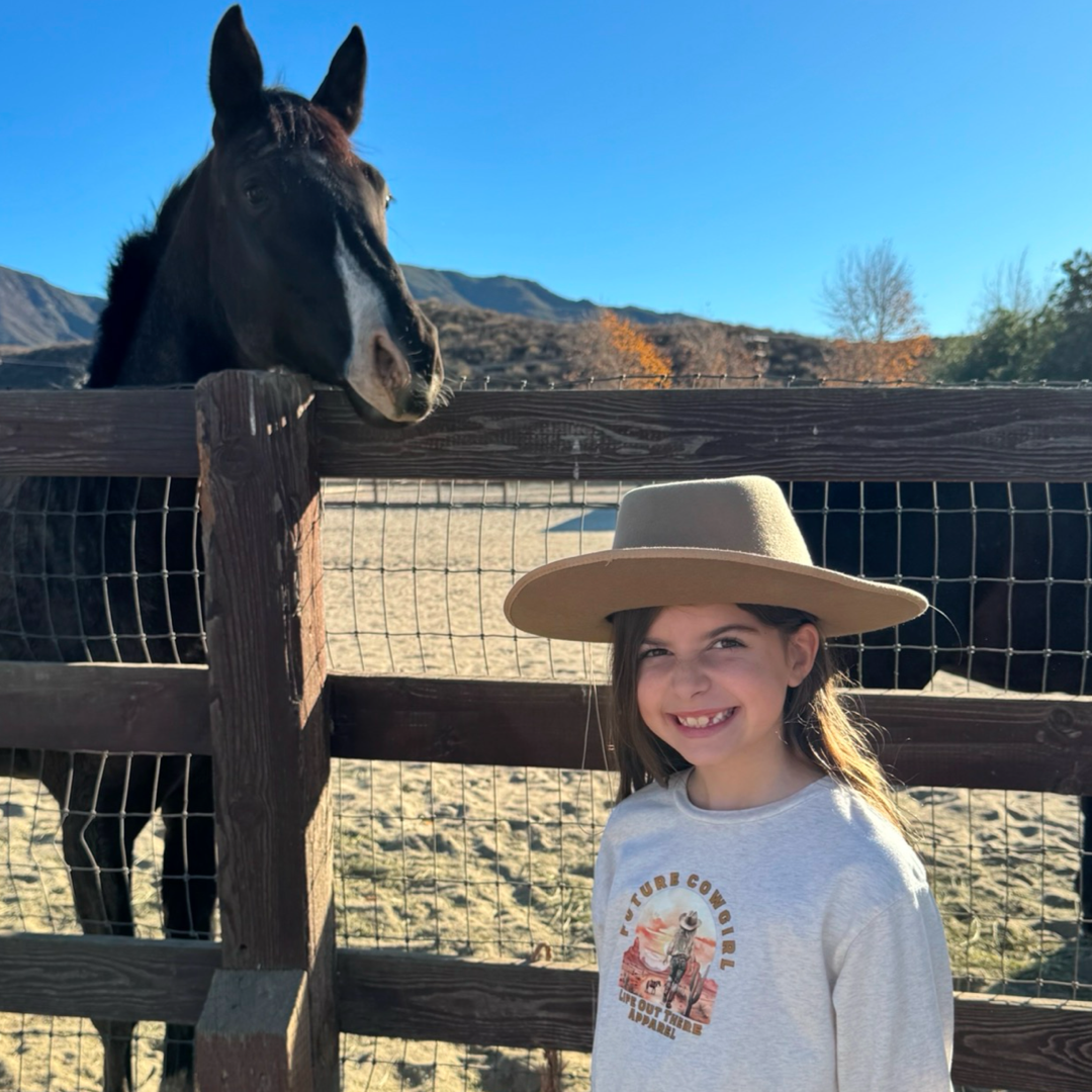 A smiling future cowgirl wearing a Life Out There Apparel Future Cowgirl T-shirt in natural beige stands near a brown horse by a wooden fence, with distant mountains under a clear blue sky.