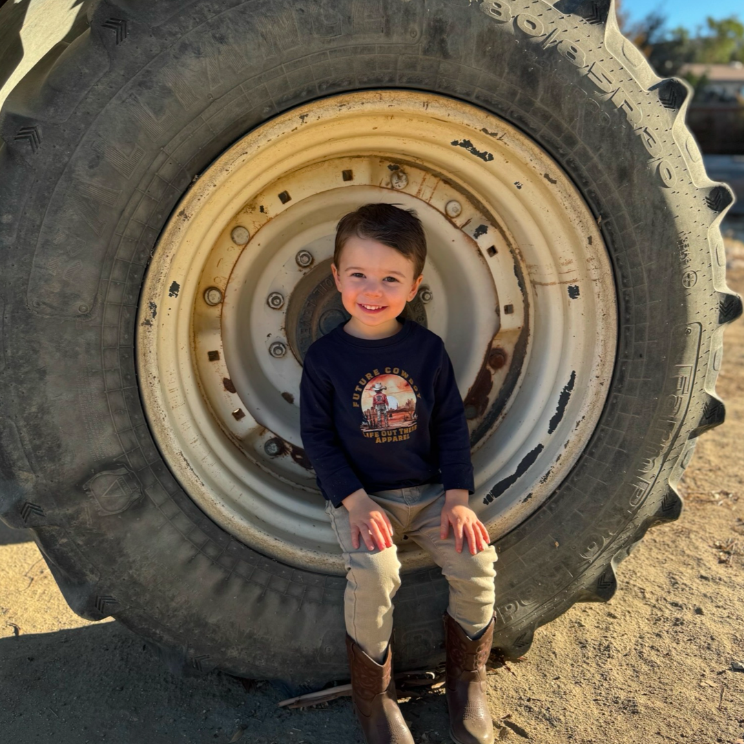 A young child with brown hair sits smiling in front of a large tractor tire, embodying a future cowboy. The child wears a navy blue Boys Youth Future Cowboy T Shirt from Life Out There Apparel, light pants, and sturdy brown boots. The sun casts a shadow, evoking classic country music vibes.