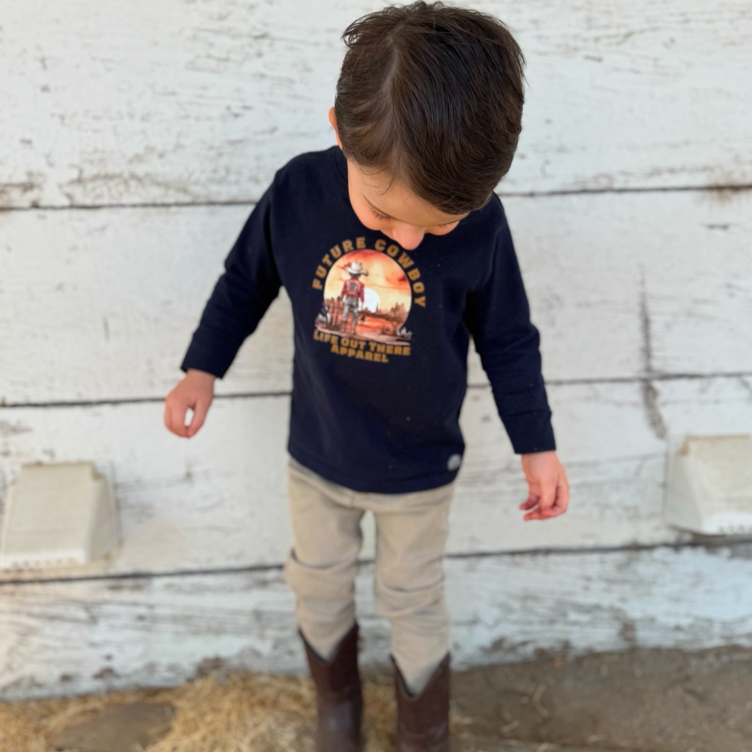 A young boy in a Life Out There Apparel Boys Youth Future Cowboy tee (available in navy, black, and red, long or short sleeve) and boots stands against a white wooden background, evoking the spirit of country music as he gazes downwards.