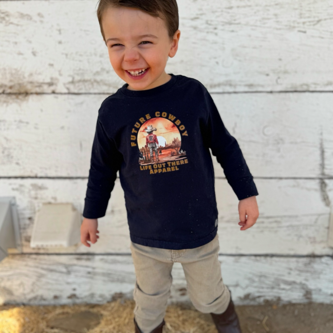 A child with short dark hair and a joyful smile stands against a white wooden background, wearing Life Out There Apparels navy Future Cowboy T-shirt, light-colored pants, and brown boots.