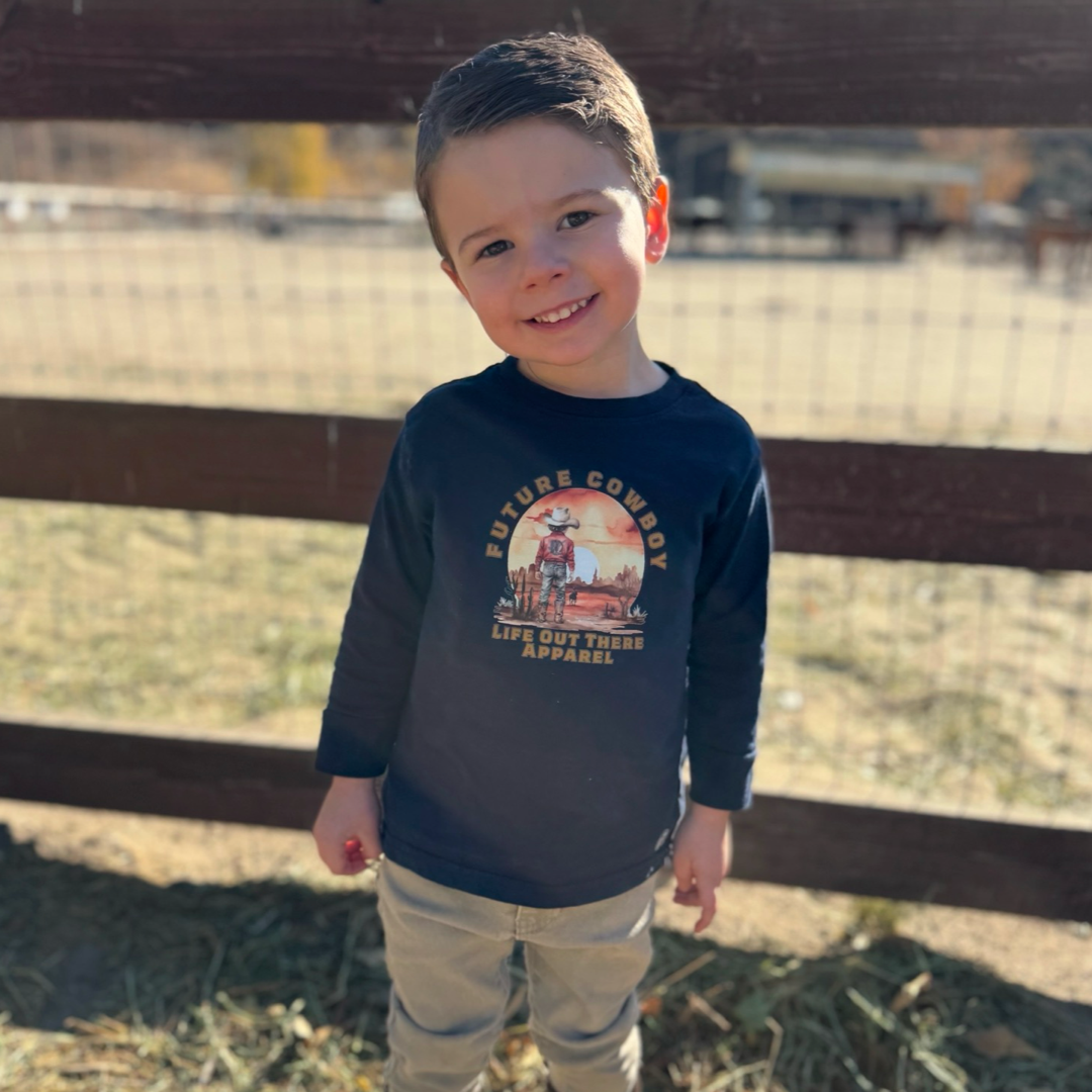 A young boy with short brown hair stands outside, wearing a navy Future Cowboy tee by Life Out There Apparel and beige pants. He slightly smiles, leaning on a wooden fence. The sun casts a warm glow, evoking country charm, with blurred trees in the background.