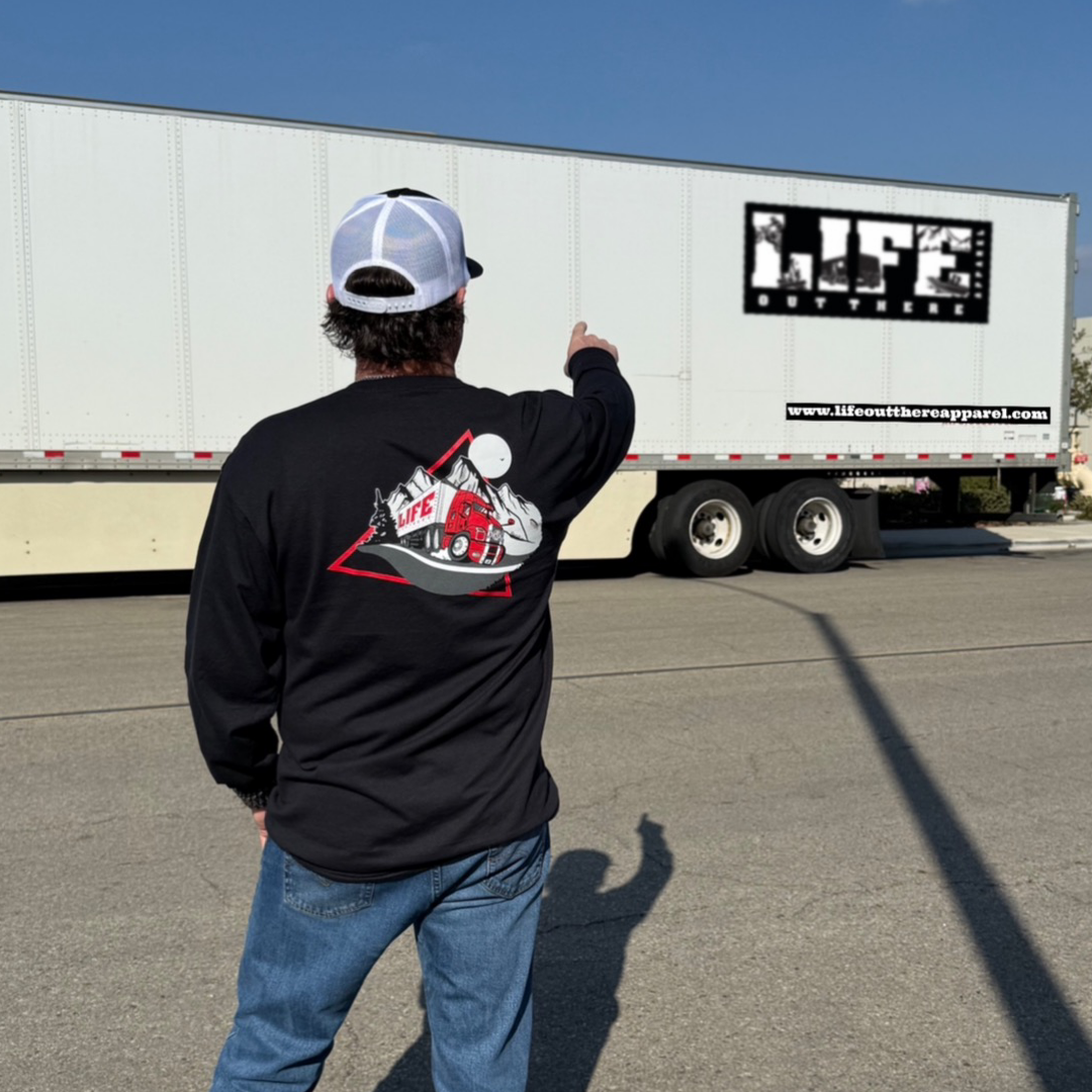 A person wearing a Life Out There Apparel 10-4 Good Buddy black long sleeve tee and a stylish black and white cap points at a large white truck trailer on a sunny street. The trailer displays the text LIFE with a website URL, capturing the spirit of truck drivers.