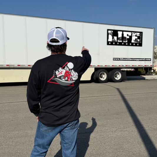 A man in jeans and the black "10-4 Good Buddy" long sleeve tee by Life Out There Apparel stands on the street, pointing at a white LIFE semi-trailer. The shirt showcases mountains, a road, and "LIFE"—ideal for truck drivers.