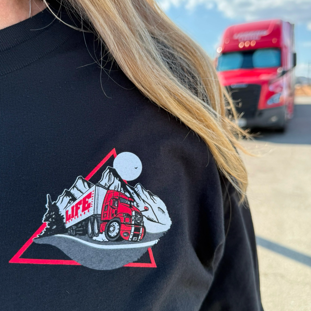 A person wearing Life Out There Apparels Unisex - 10-4 Good Buddy - Long Sleeve Tee - Black, featuring a red truck and mountain design with LIFE on the truck. A parked red truck in the background under a clear sky emphasizes this designs appeal to truck drivers.