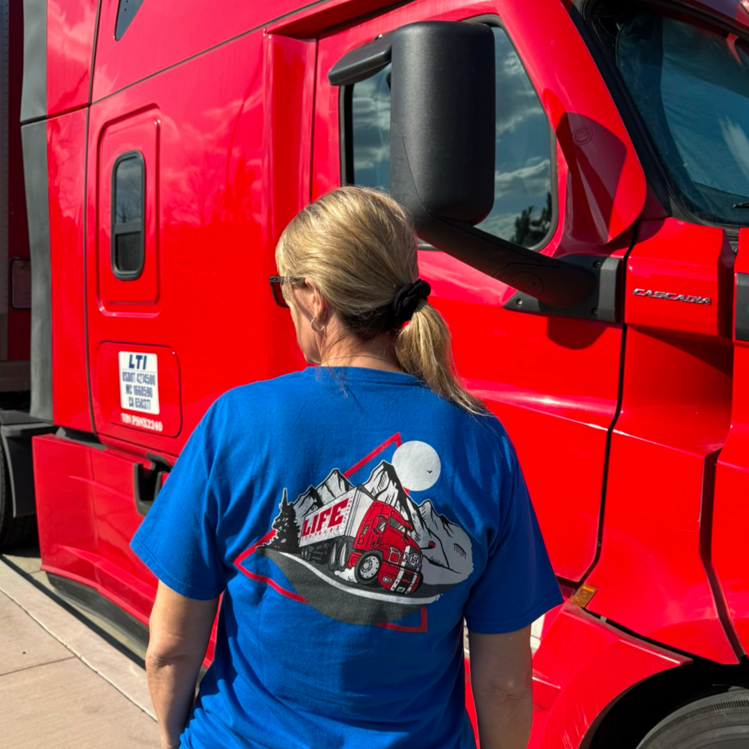 A blonde-haired person in a ponytail wears the Life Out There Apparels blue Unisex - 10-4 Good Buddy Tee - Royal, made from 50% cotton, with a red truck graphic. They stand confidently before a red truck with a logo, embodying proud truck drivers spirit under partly cloudy skies.