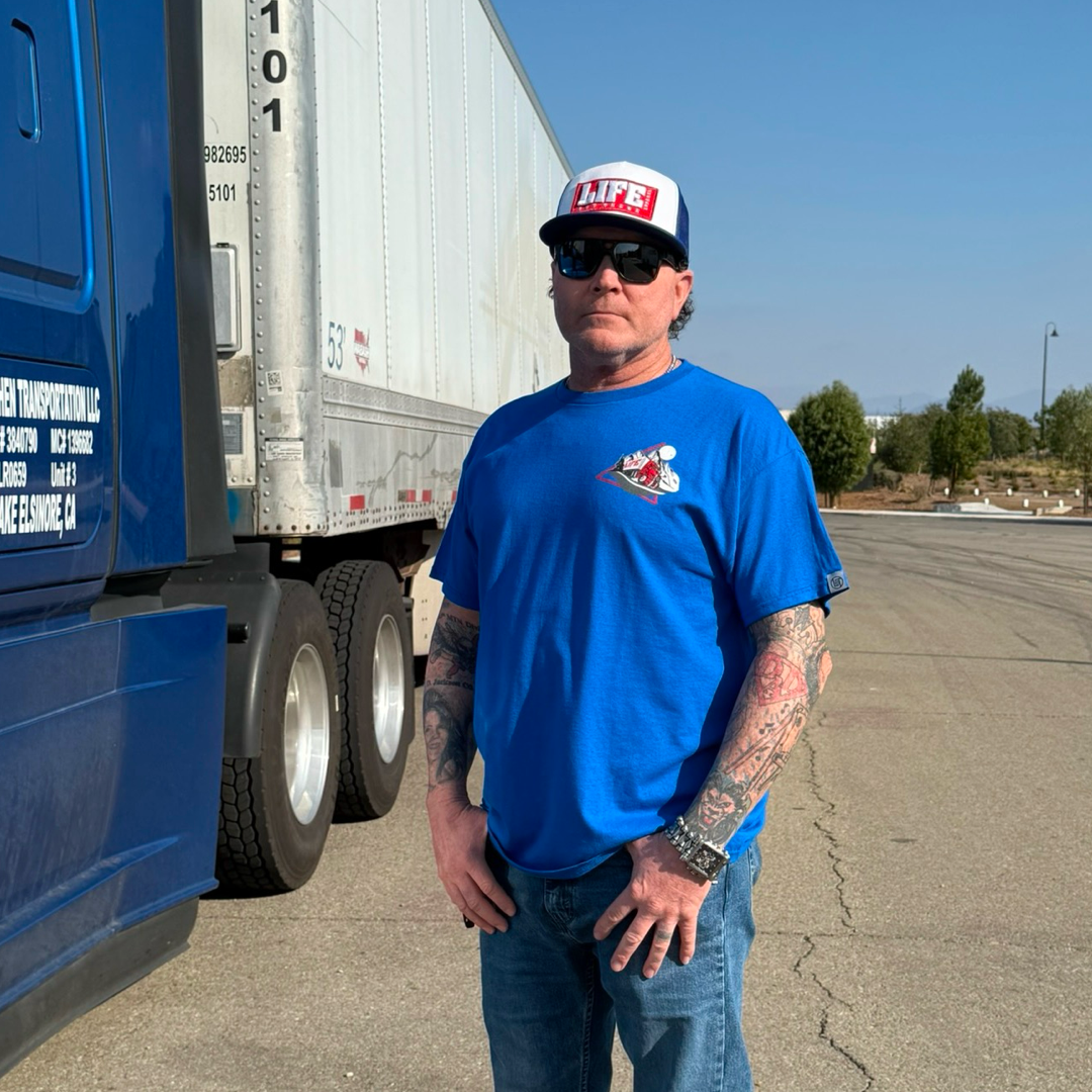 A man confidently stands beside a large blue truck wearing Life Out There Apparels Unisex - 10-4 Good Buddy Tee in Royal. With sunglasses, vibrant tattoos, and a cap, his look is classic among truck drivers. Trees in the background add a natural touch under the sunny sky.