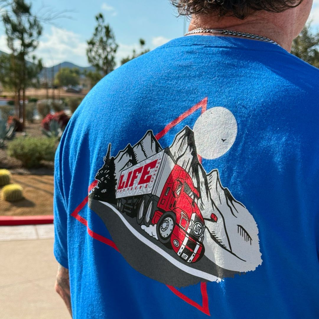 Wearing the Unisex - 10-4 Good Buddy Tee in royal blue by Life Out There Apparel, this 50% cotton T-shirt features a red semi-truck driving in front of mountains and a full moon. Perfect for truck drivers, it boldly displays LIFE above the truck.