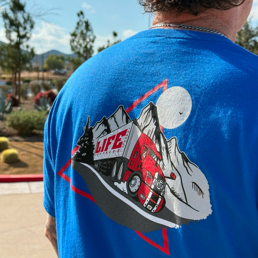 A person wears the 10-4 Good Buddy Tee - Royal by Life Out There Apparel, featuring a red semi-truck, mountains, trees, and a full moon with "LIFE" on the truck; the scene is outdoors with plants and trees in the background.