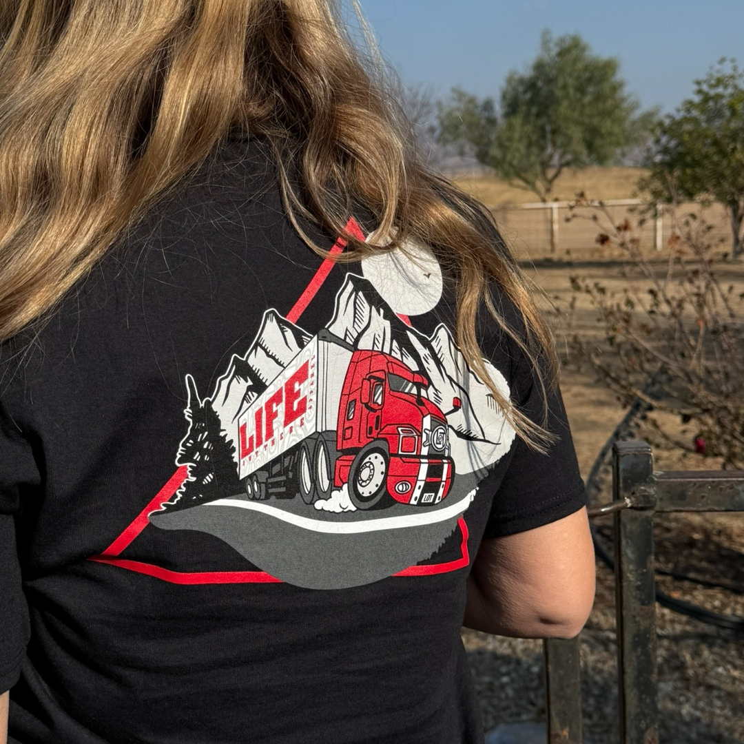 A person with long hair wears the Life Out There Apparels Unisex - 10-4 Good Buddy Tee in black, featuring a red truck against rugged mountains and Life printed on it. A fenced outdoor area with trees and grass complements this unique piece of apparel, capturing the spirit of truck drivers.