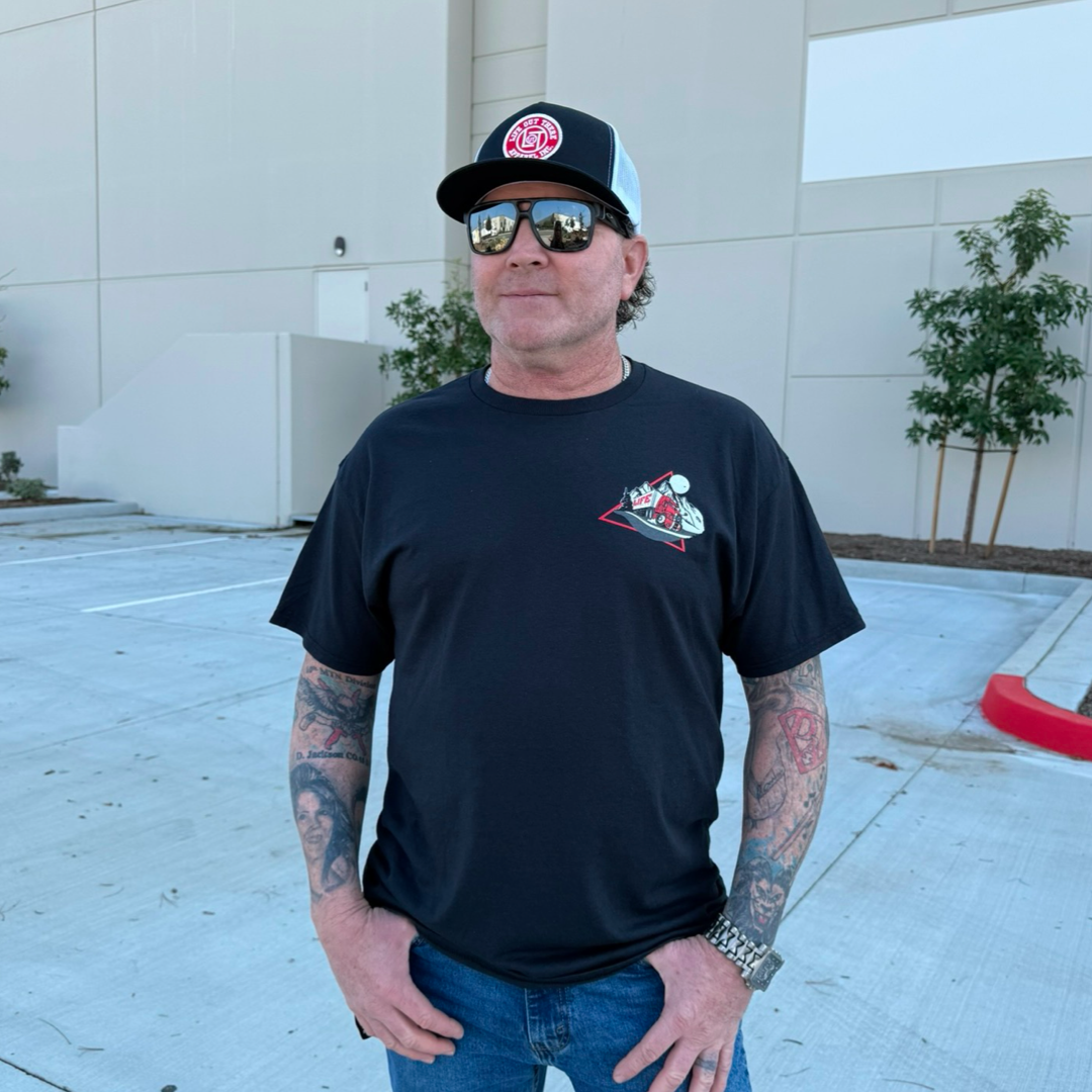 In a parking lot, someone dons sunglasses, a black cap, and Life Out There Apparel’s Unisex 10-4 Good Buddy Tee in black, featuring trucker-inspired graphics. Tattoos cover their arms alongside a watch. A beige building with green shrubs stands behind them, contrasting with their bold attire.