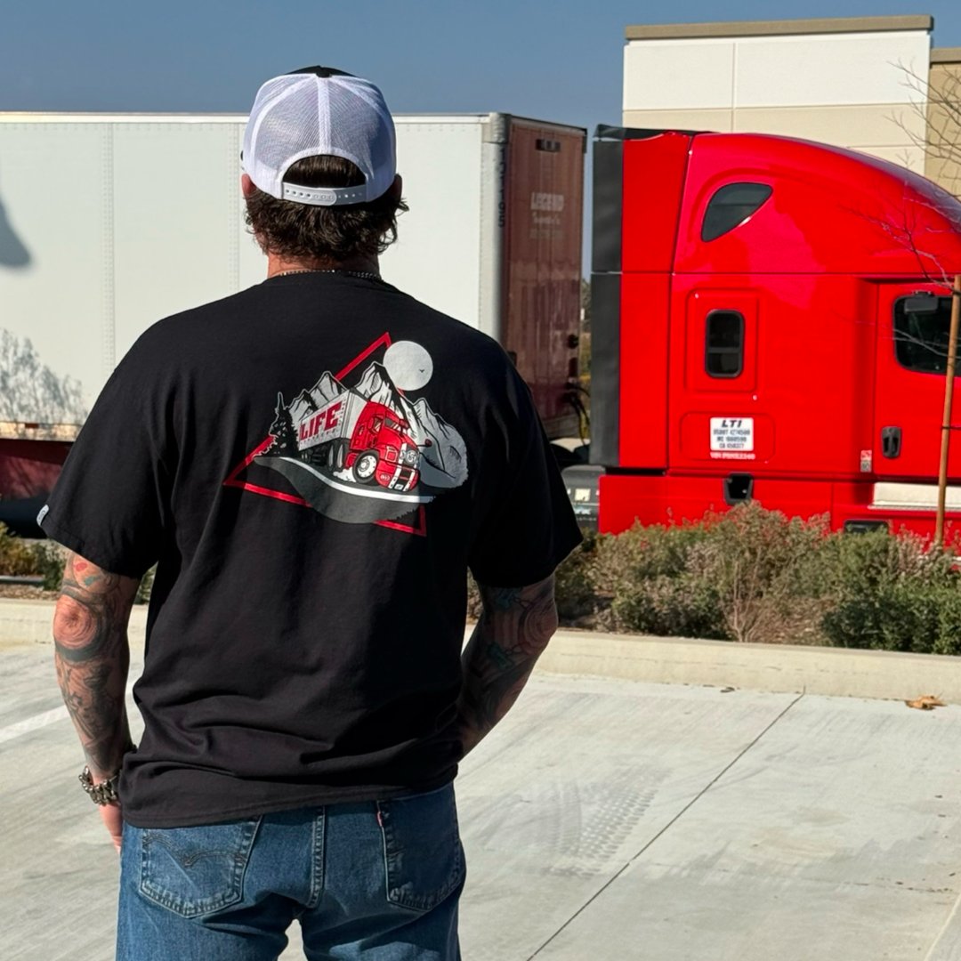 A tattooed person faces away, wearing a Life Out There Apparel Unisex - 10-4 Good Buddy Tee in black with a red truck design. A nearby red semi-truck highlights pure transportation under a clear, sunny sky.