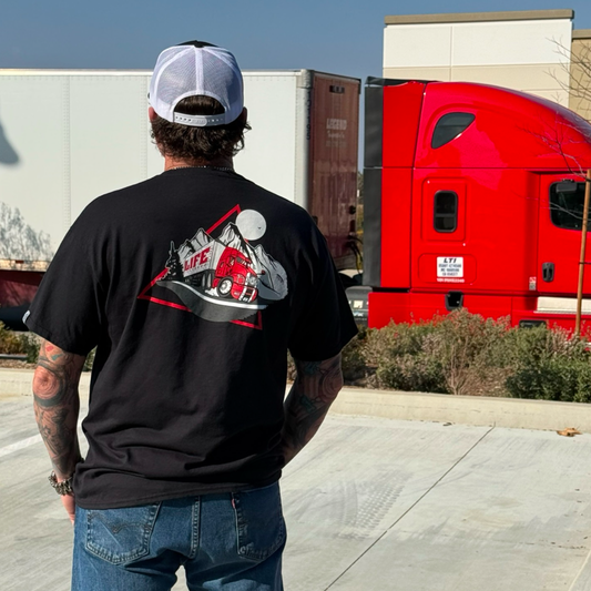 A tattooed person stands outdoors facing away, wearing the Life Out There Apparel 10-4 Good Buddy Tee - Black. The shirt’s graphic shows a red semi-truck and mountains; a real red semi-truck is parked in the background—classic trucker style.