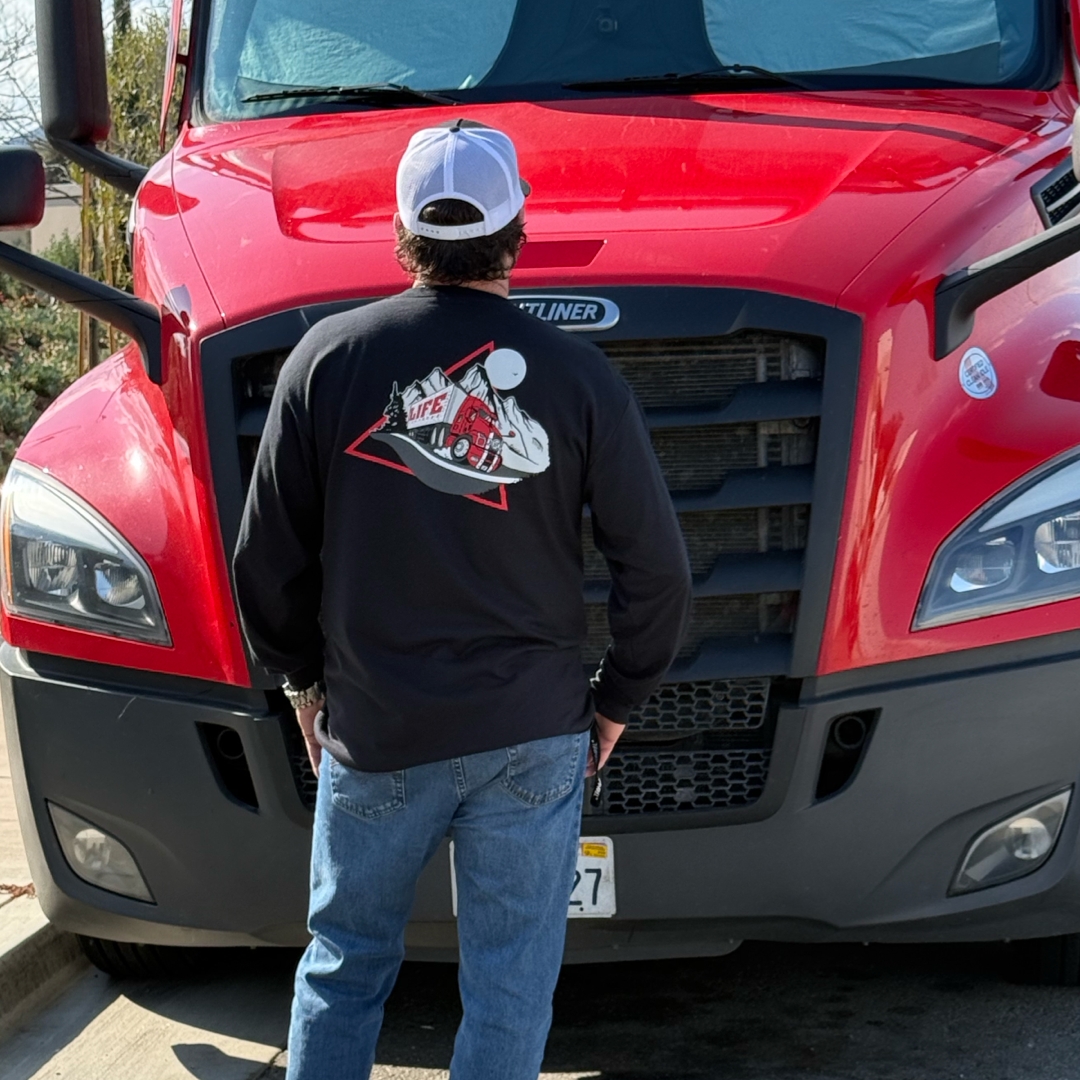 Wearing Life Out There Apparels Unisex 10-4 Good Buddy Black Long Sleeve Tee and a white cap, a man confidently stands in front of a parked red truck with a tall grille, capturing the rugged essence of truck drivers.