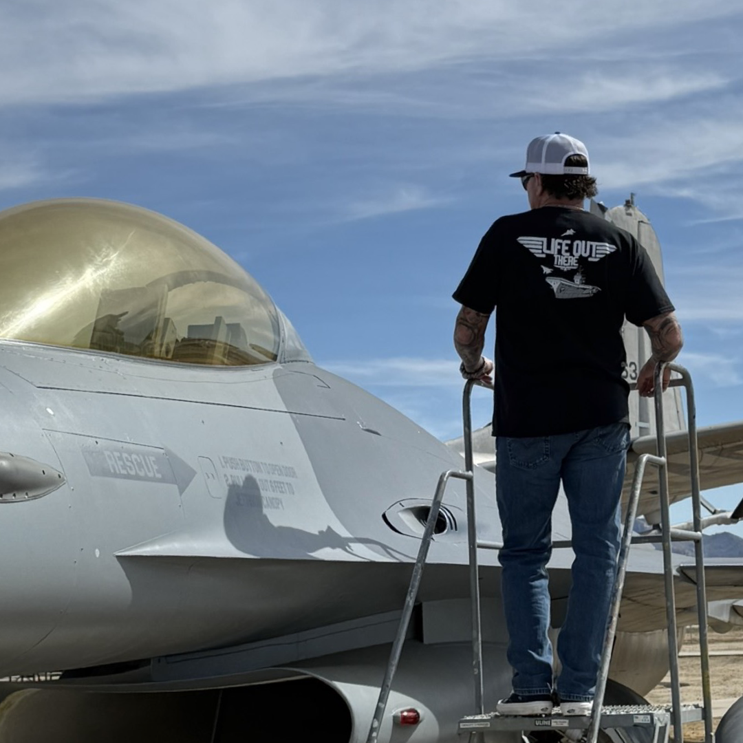 A person wearing Life Out There Apparels Unisex The Hard Deck Tee in black and a TOPGUN-styled cap stands on a stepladder next to a grey fighter jet, gazing at its cockpit against a backdrop of partly cloudy skies, celebrating iconic US military aviation.