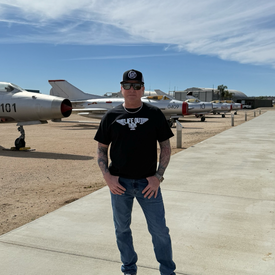 A man wearing a black Unisex - The Hard Deck Tee by Life Out There Apparel and jeans stands on a concrete path with his hands in pockets. With sunglasses and a baseball cap, he admires vintage fighter jets behind him, evoking a classic TOPGUN scene beneath the clear blue sky.