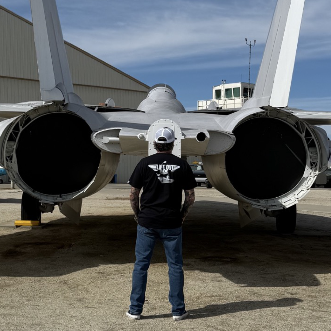 A person wearing the Life Out There Apparels Unisex - The Hard Deck Tee in black and a white cap stands on concrete, facing the rear of a fighter jet. Its large engines and tail fins hint at TOPGUN prowess, while a building looms in the background under a clear blue sky.