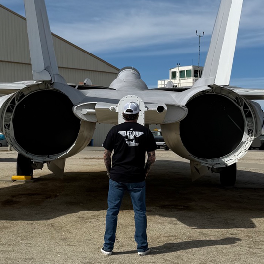 Wearing the Life Out There Apparel Hard Deck Tee in black, a person pairs it with jeans and a white cap while standing behind a US Navy fighter jet with dual engine exhausts, evoking TOPGUN vibes amid hangar buildings.