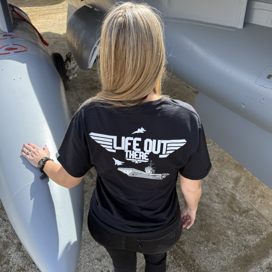 A person with blonde hair, wearing the Life Out There Apparels Unisex Hard Deck Tee in black, stands facing a large fighter jet. The T-shirt features a white graphic and the text LIFE OUT THERE, as their hand rests on the aircraft, capturing TOPGUNs daring spirit.