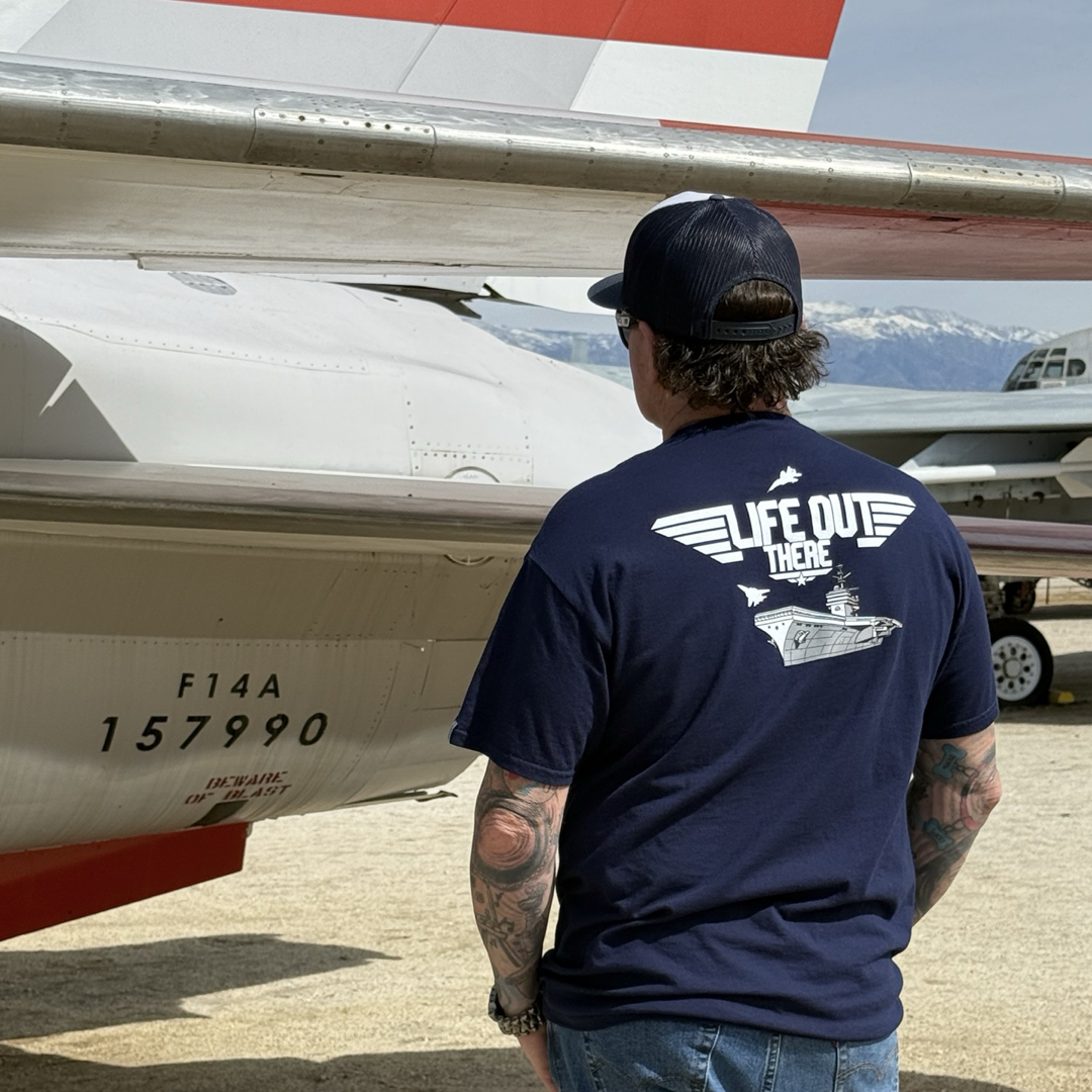 A man wearing a Life Out There Apparels Unisex The Hard Deck Tee in navy stands under an F14A aircrafts wing. The outdoor scene, with mountains and clear skies, evokes TOPGUN MAVERICK vibes.