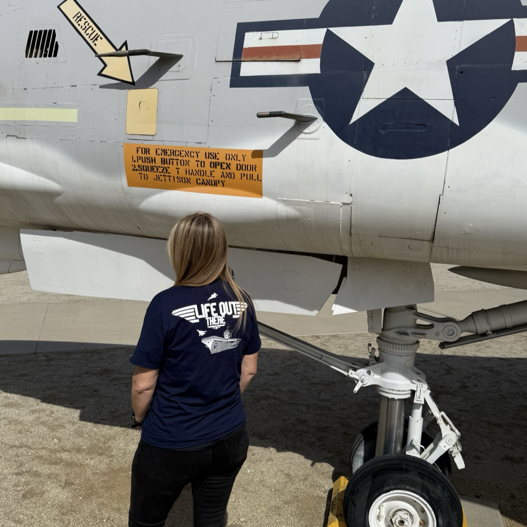 A person wearing a navy The Hard Deck Tee by Life Out There Apparel stands beside a US military aircraft, facing an emergency instruction panel. The aircraft displays a star insignia with arrows pointing to the panel, reminiscent of TOPGUN MAVERICK. Sunlight casts shadows on the ground.