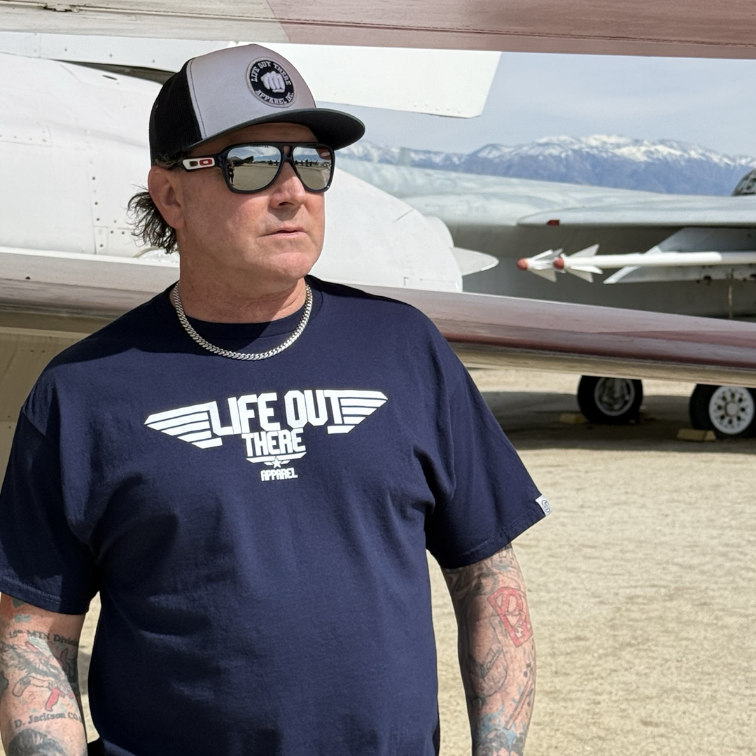 A man in a navy The Hard Deck Tee by Life Out There Apparel, sunglasses, and baseball cap stands beside an aircraft with a TOPGUN MAVERICK vibe. His tattooed arms and chain necklace catch the light against snow-capped mountains and another plane in the stunning backdrop.