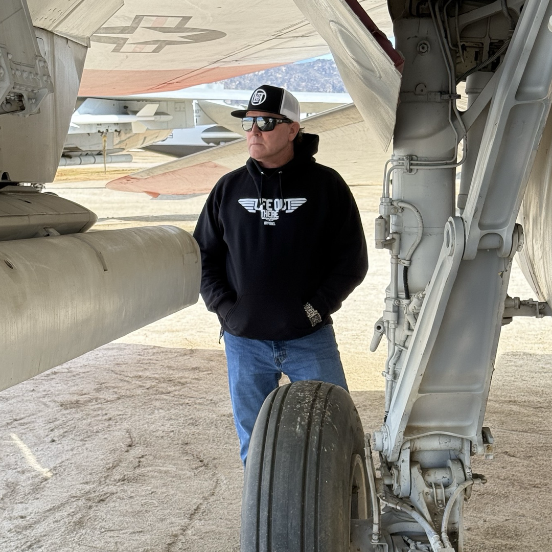 A person in a Life Out There Apparel Unisex - The Hard Deck Hoodie - Black, blue jeans, sunglasses, and a baseball cap stands under a large aircrafts wing. Fighter jets create a striking backdrop in this outdoor scene reminiscent of TOPGUN.