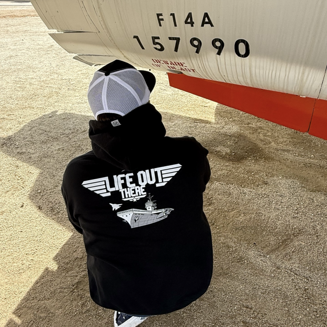 Wearing a black The Hard Deck Hoodie by Life Out There Apparel featuring an aircraft carrier and LIFE OUT THERE, a person in a white cap crouches beside an F14A jet on sandy ground, capturing the spirit of TOPGUN and evoking US Military air adventures.