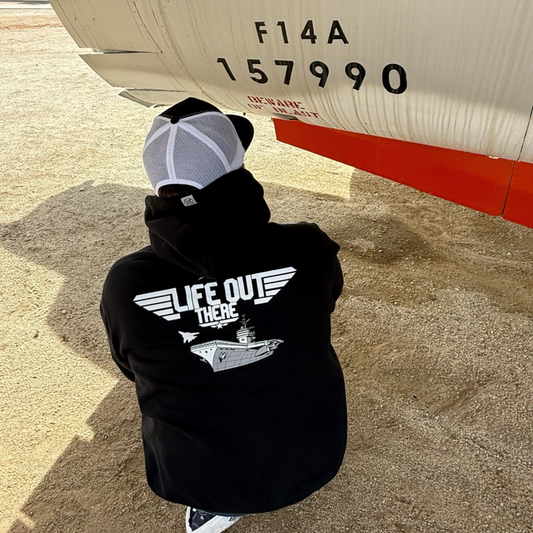 A person in The Hard Deck Hoodie - Black by Life Out There Apparel crouches near the nose of a white F14A 157990 fighter jet, wearing US Navy-inspired gear on sandy ground.