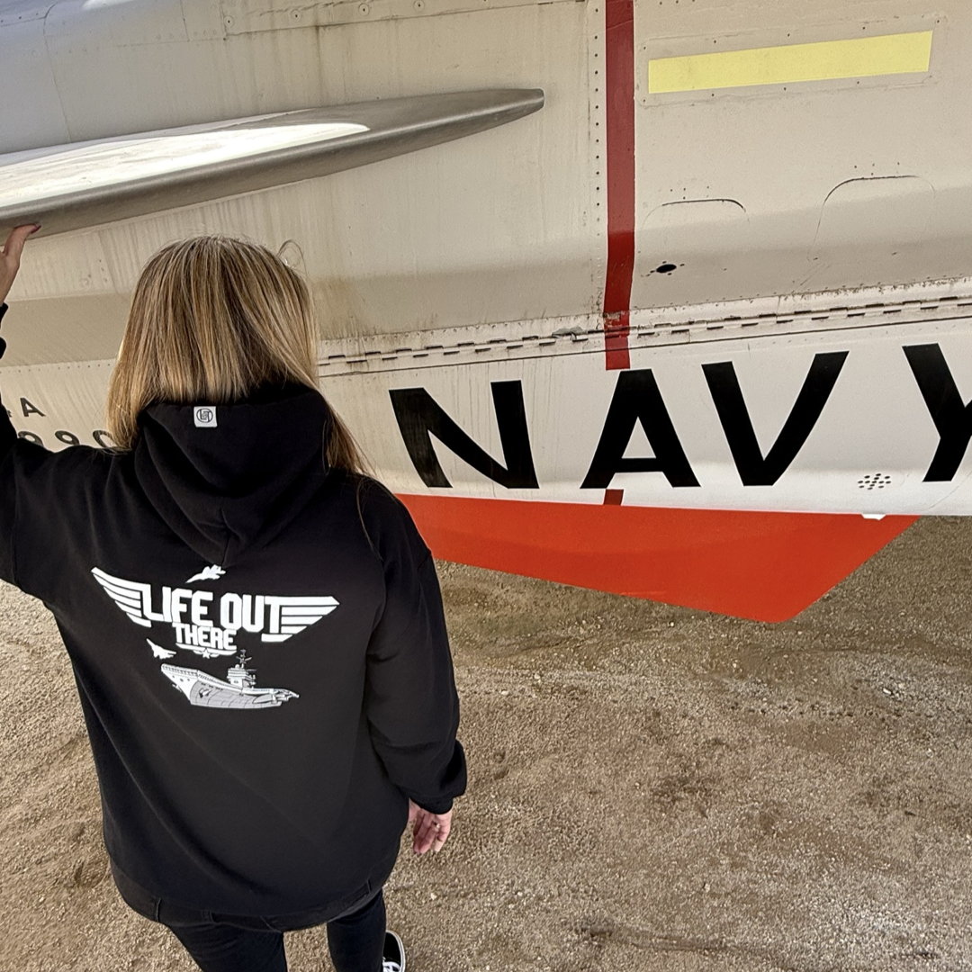 A person wearing the Unisex - The Hard Deck Hoodie - Black by Life Out There Apparel stands beside a vintage navy aircraft, reminiscent of TOPGUN days, with a mostly white body, red stripe, and prominent NAVY, reflecting its US Military fighter jet heritage.