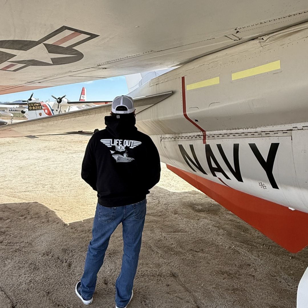 A person in a Life Out There Apparel Unisex - The Hard Deck Hoodie - Black and jeans stands under a vintage military aircraft wing with NAVY on it, gazing at parked fighter jets like a TOPGUN scene in an outdoor museum.