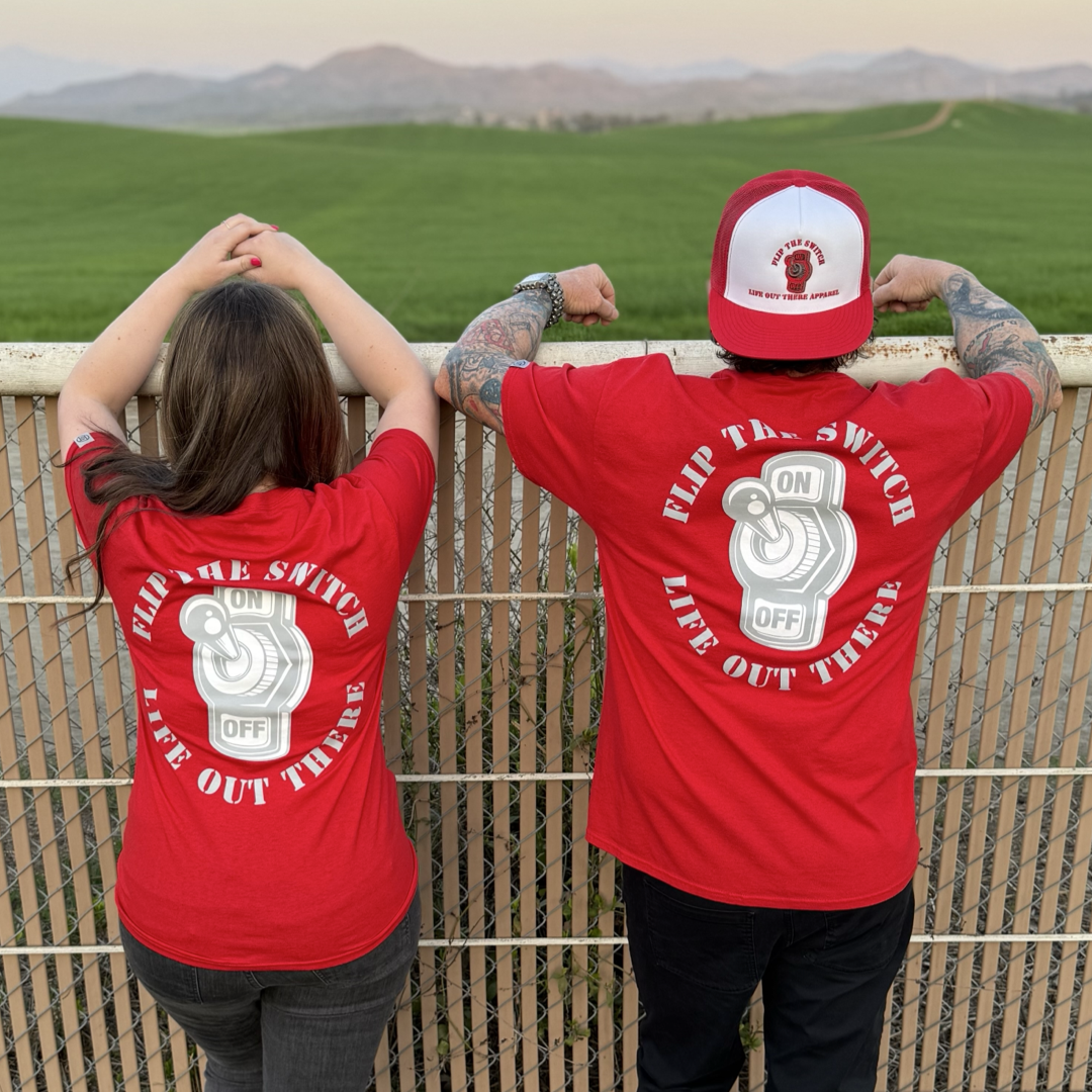 Two people in matching red Unisex Flip the Switch tees and caps stand by a fence overlooking lush fields and mountains. Their shirts feature a light switch graphic with ON and OFF, and the text Flip the Switch, Live Out There, marking Life Out There Apparels anniversary launch.